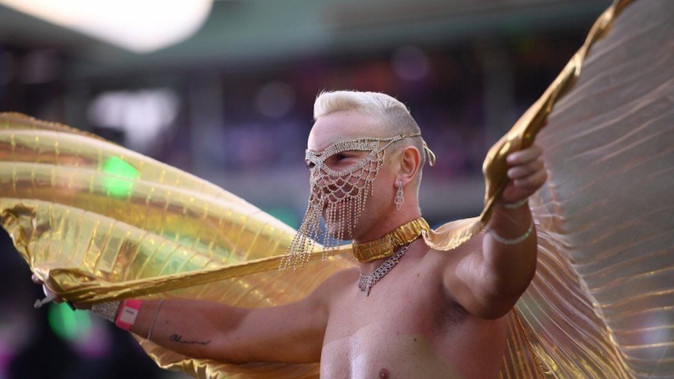 A man wears a jewelled gold face mask and large gold wings during parade