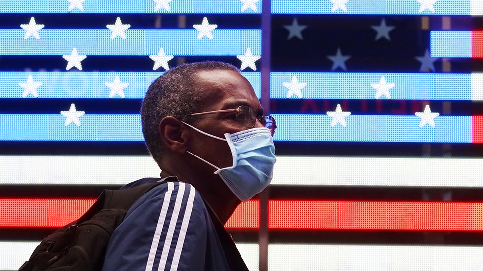 A person wearing a face mask walks past lights displaying the US flag in Times Square, New York - 22 October 2020