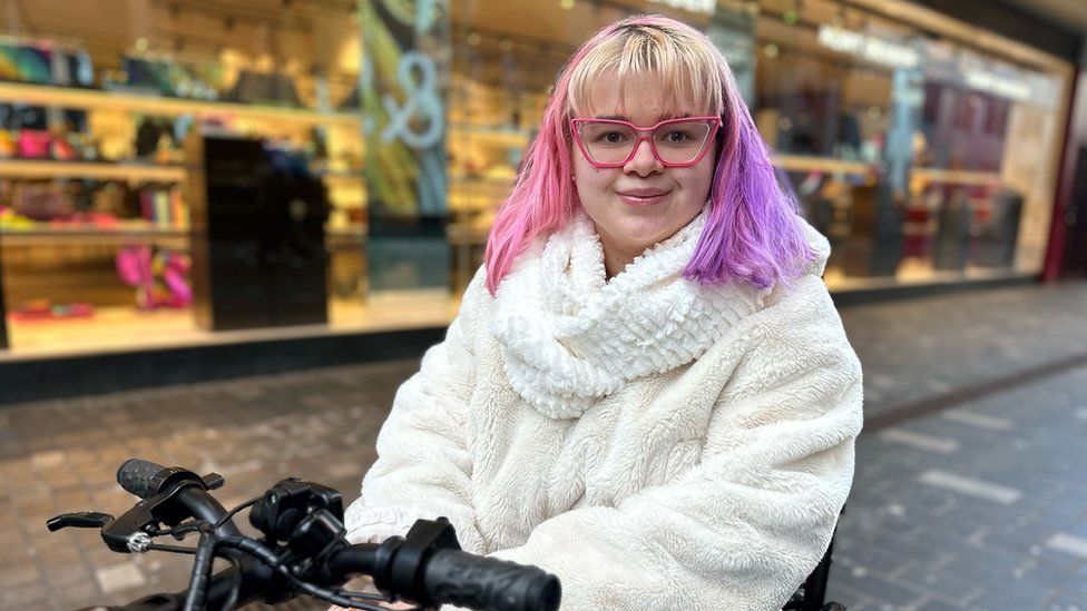 Sophia Dunn. Sophia is a 19-year-old white woman. She has hair dyed pink on one side and purple on the other with a blonde fringe. She has pink rimmed glasses framing her brown eyes and is smiling at the camera. She wears a fluffy cream jumper with matching scarf and is on a mobility scooter she uses. Sophia is pictured outside a clothing shop in Liverpool