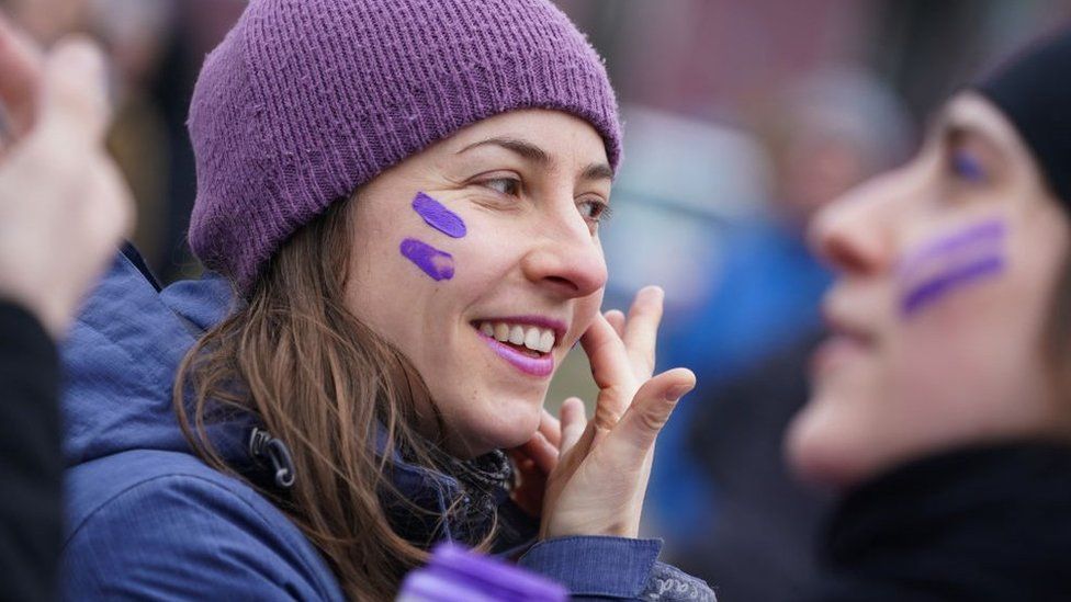 A woman wearing a purple hat puts purple face paint on her cheeks at march to mark International Women's Day