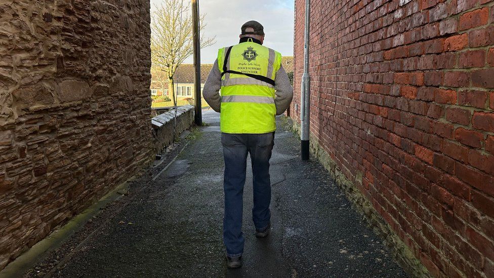A man in a high vis jacket walks away from the camera down an alleyway between two brick walls