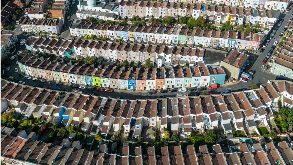 An aerial view of terraced streets in Bristol, UK. Seven streets of homes, arranged in a uniform pattern, are broken up only by roads linking them. The streets are colourful, with houses on each painted different colours. There are a range of neutral and pastel shades on display, with one lime green frontage drawing particular attention. The angle of the plentiful parked cars tells the viewer that the streets are on a hill. It's a heavily residential area, with only one building - a white, domed mosque - towards the rear of the photo that appears to serve another purpose.