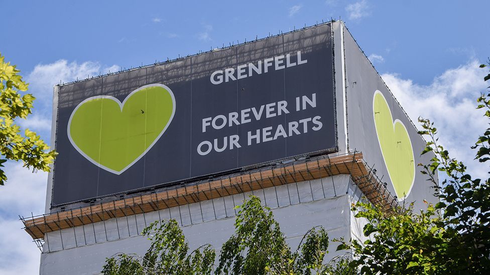 A billboard on the side of Grenfell Tower reads 'Grenfell, forever in our hearts' next to a big image of a green heart, with trees in the foreground. Picture taken on the seventh anniversary of the Grenfell disaster.