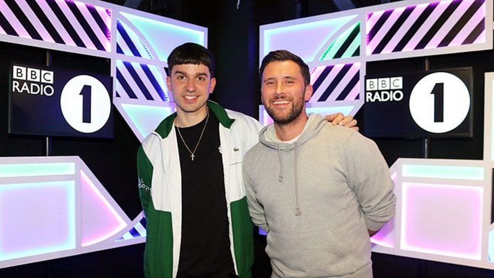 Michael Bibi with Danny Howard in the Radio 1 studios. Michael is wearing a black T-shirt with a green and white jacket over it and Danny is wear a beige hoodie. Both are smiling for the camera.