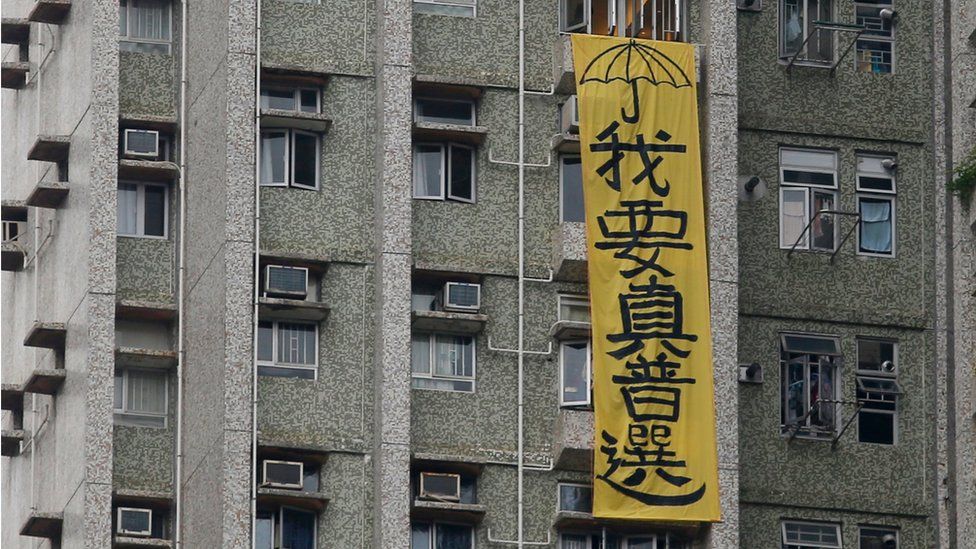 A vertically displayed yellow banner, with black text, hanging on the side of a block of flats, which reads "I need real universal suffrage", in Hong Kong on 19 May