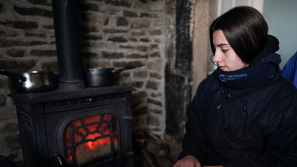 Молодая женщина в пальто сидит у дровяной печи во время отключения электроэнергии