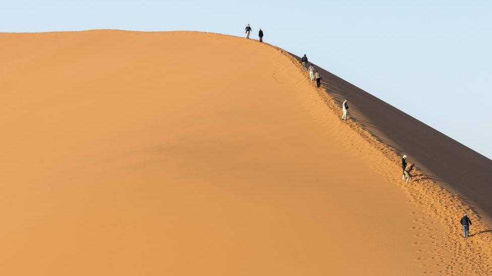 Tourists Climbing Along An Edge Of A Huge Sand Dune At Namib Desert. Namibia.