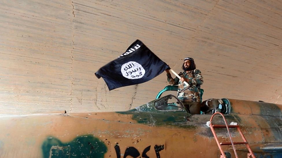 Islamic State fighter (ISIS; ISIL) waving a flag while standing on captured government fighter jet in Raqqa, Syria 2015