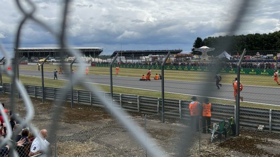 View from behind a wire fence of part of the Silverstone circuit. Several people wearing orange are running, standing or sitting on the track. Others, possibly marshals, are running towards them