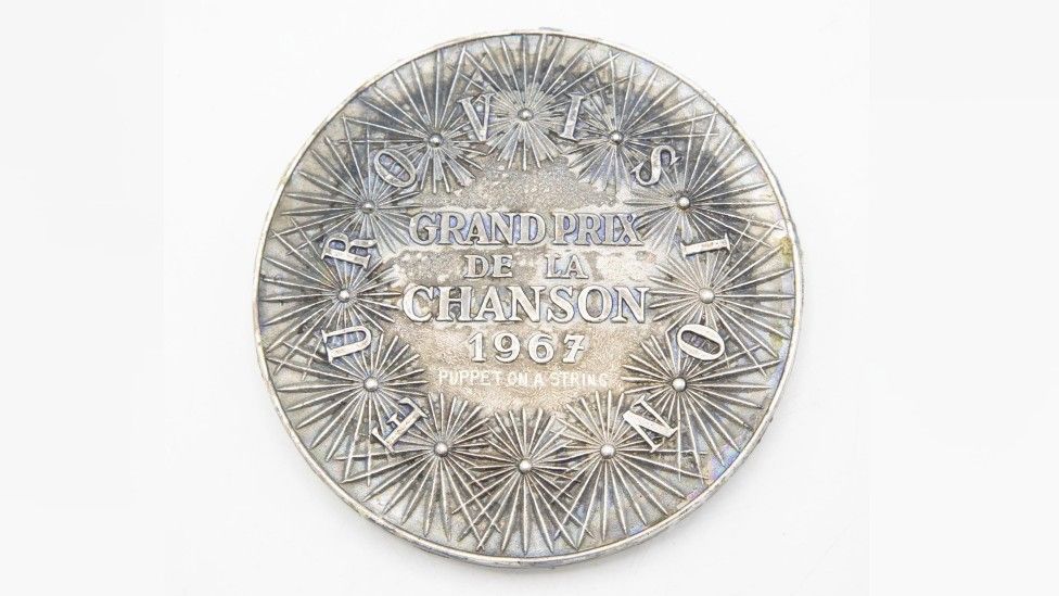Reverse of Medal. A series of spiky shaped flowers adorn the smooth edges of the medal, with the words Eurovision Grand Prix de la Chanson 1967, Puppet on a String written on it.