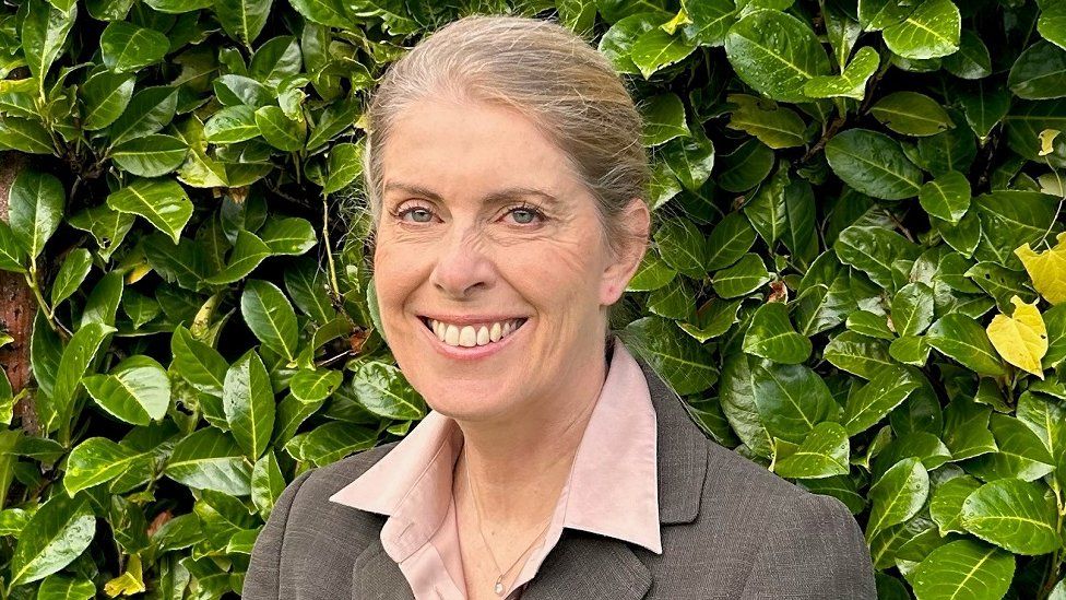 Woman with short fair hair tied back, wearing a grey jacket and standing in front of a hedge