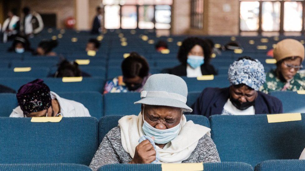 Women with emotional expressions at a funeral. They are sitting in blue pews and appear to be socially distanced. April