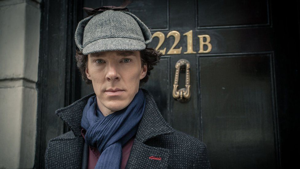 Benedict Cumberbatch dressed in a deerstalker outside 221B door.