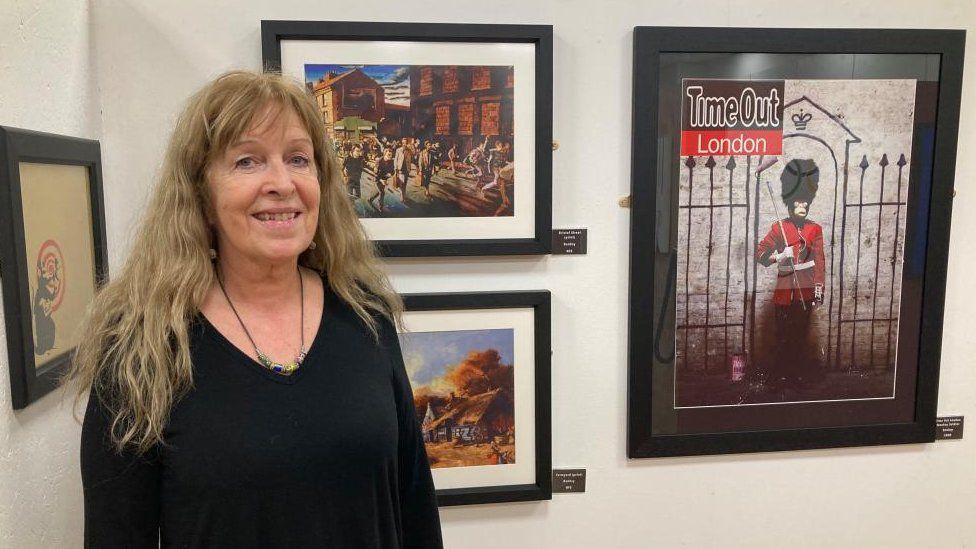 Woman with long light brow hair wearing black stands in front of pictures on a wall. One shows a guardsman.