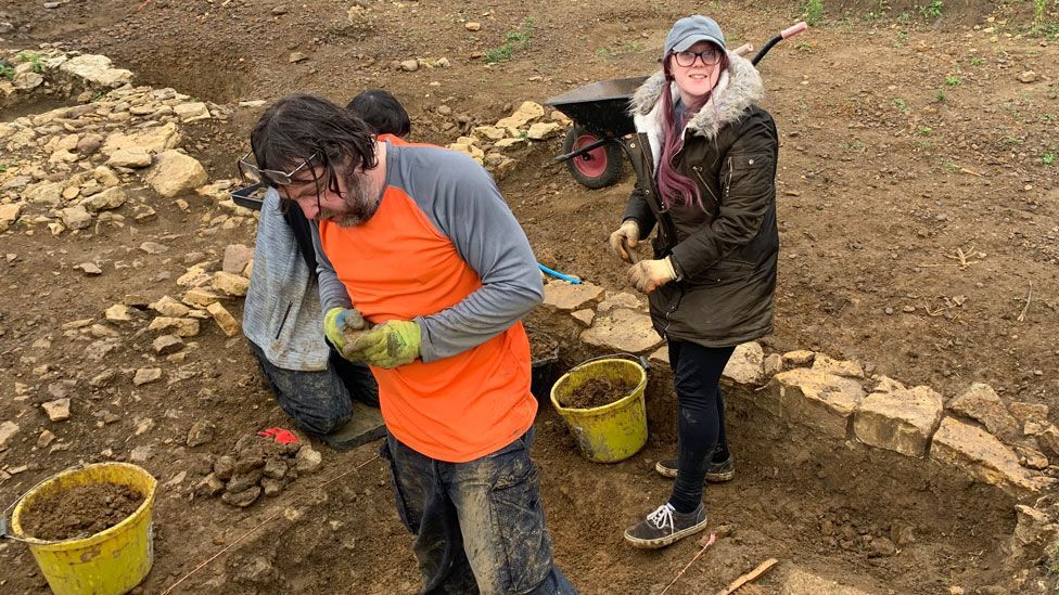 A man and a woman standing in a trench edged in stone during an excavation, quite muddy, and with yellow buckets