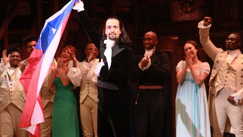 Lin-Manuel Miranda standing on-stage at the end of a performance of Hamilton, holding the Puerto Rico flag
