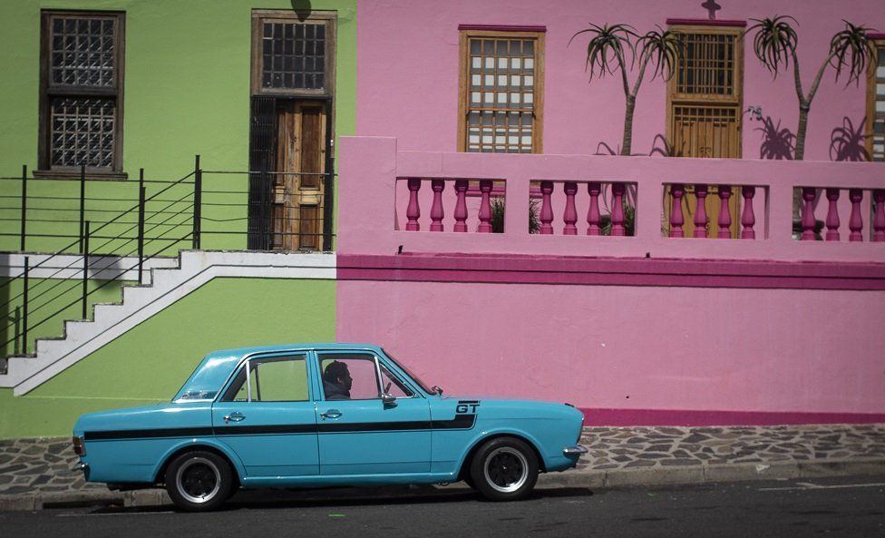 A blue car parks in front of pink and green houses.