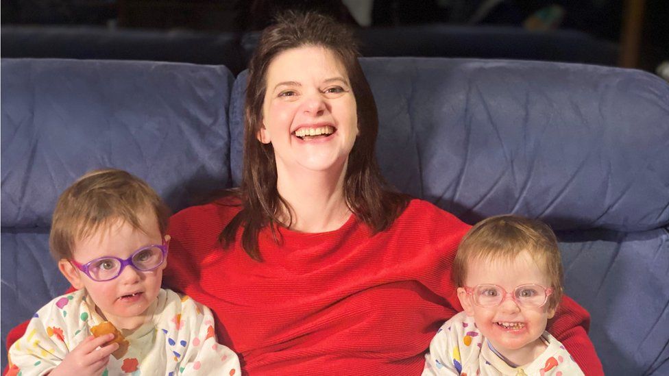 A white woman in a red jumper with brown hair smiles at the camera with her two toddler daughters sat either side of her, both girls wear colourful glasses