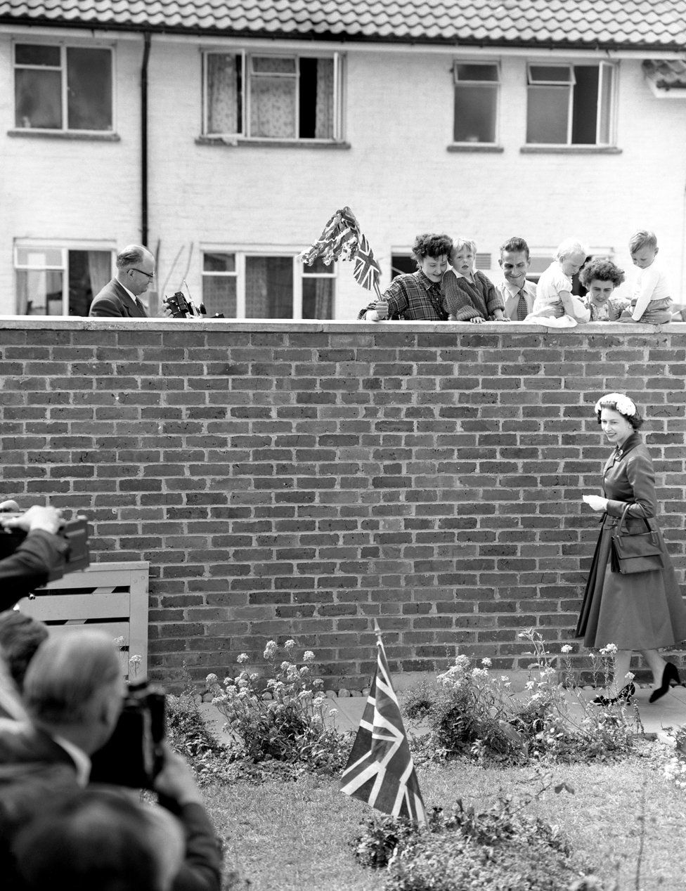 A Union Jack planted in the garden caught the Queen's attention, but she is the object of the neighbours' gaze. They watched as she arrived to visit the home of Mr and Mrs Eddie Hammond at Crawley New Town, Sussex after opening Gatwick Airport. 1958