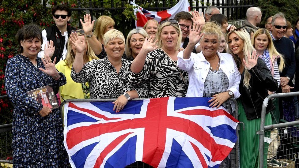A group of women standing behind a Union flag outside Hillsborough Castle wave and smile after seeing the King and Camilla