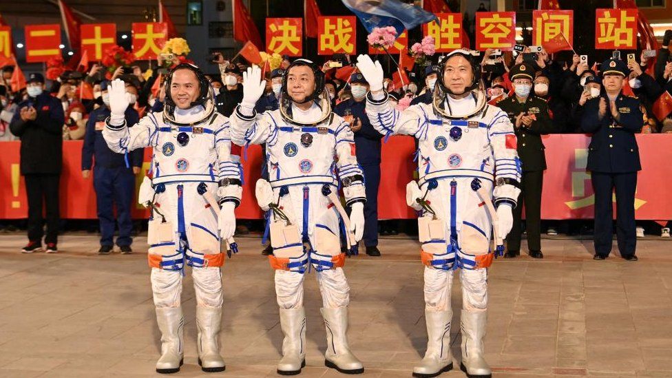 Трое астронавтов стоят в скафандрах перед китайскими официальными лицами и машут рукой