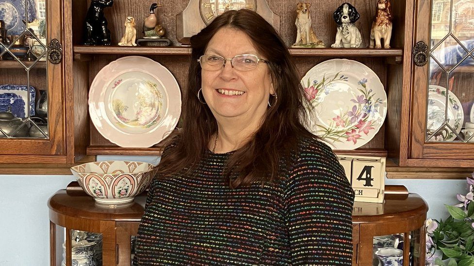 Sue Noades, with long brown hair, glasses and a multicoloured top in front of vintage ornaments