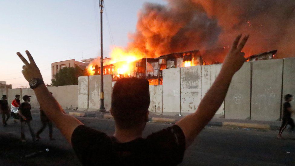 A protester stands with arms raised in front of a burning government building in Basra, 6 September 2018
