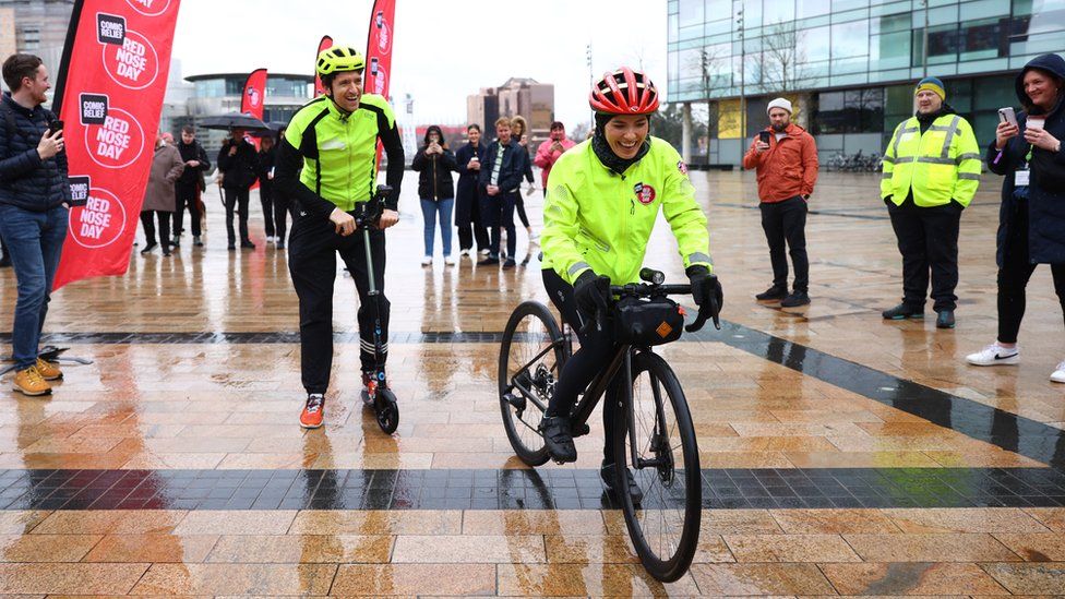A woman rides a bicycle on a rain-covered plaza. She's wearing a bright red crash helmet and a bright yellow hi-vis jacket. Behind her, a man in a similar jacket and bright yellow helmet attempts to follow on a two-wheeled child's scooter. He's too tall for the vehicle, lending the image a comedy feel. Onlookers watch the pair with amusement in the background.