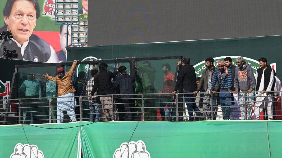 Men move into position a number of framed glass panels on a stage, with a large image of Imran Khan seen in the background