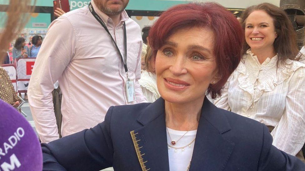 Sharon Osbourne wearing a dark blazer smiles while being interviewed while a woman over her left shoulder smiles at the camera and a man stands behind her right shoulder.