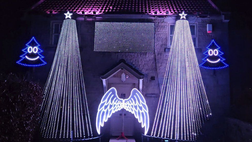 Image of the light display at Faye and Richard Dawson's home. There are two large Christmas trees made up of LED lights either side of their front door. There is also a pair of Christmas tree lights, which have smiley faces. LED Angel wings are placed on their front door.
