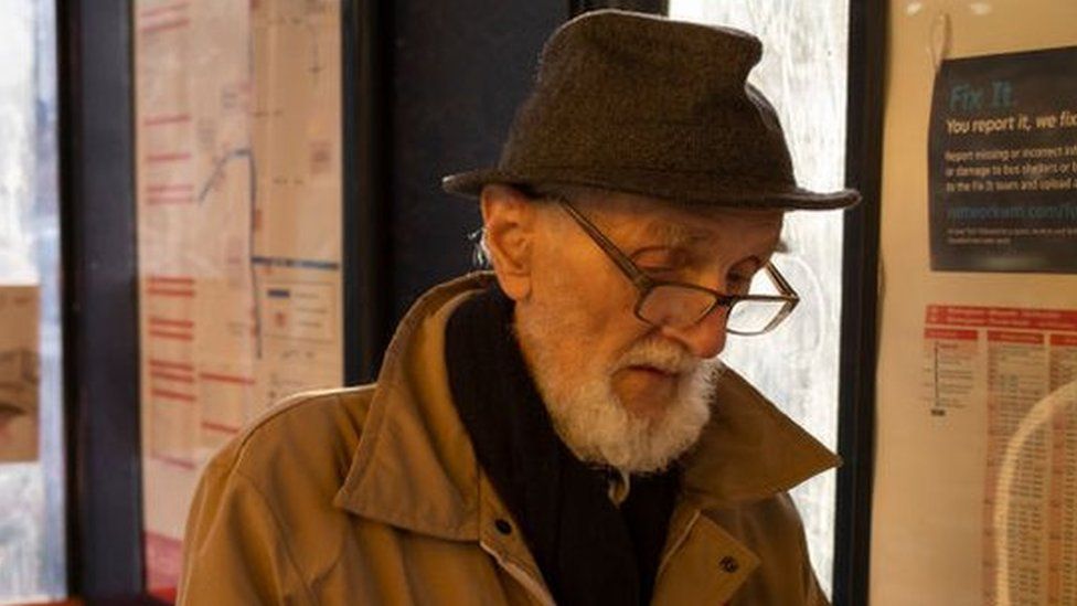 Elderly man wearing a hat and glasses reads a book at a bus stop