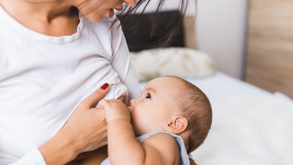 A woman breastfeeding a baby. They are both dressed in white. A bed can be seen in the background