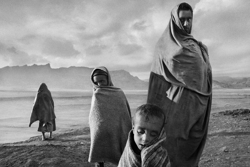 Draped in blankets to keep out the cold morning wind, refugees wait outside Korem camp. Ethiopia, 1984