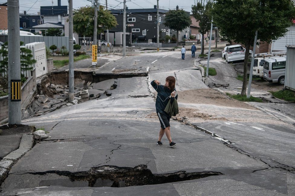 A woman takes a photograph as she walks along a road that was heavily damaged by an earthquake