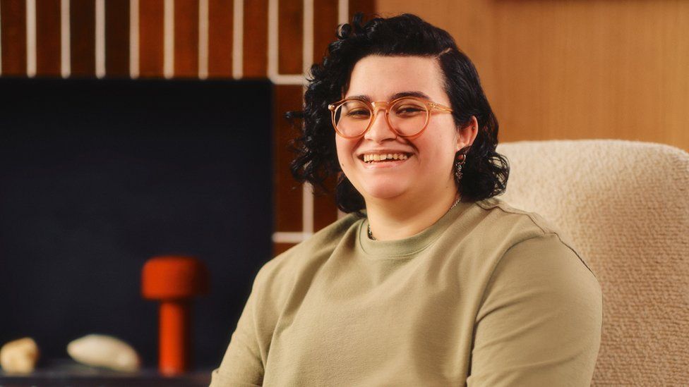 A woman with short, wavy black hair sits on a beige chair in a wood-panelled room. She wears glasses with large, opaque, round plastic frames. She's smiling, perhaps laughing.