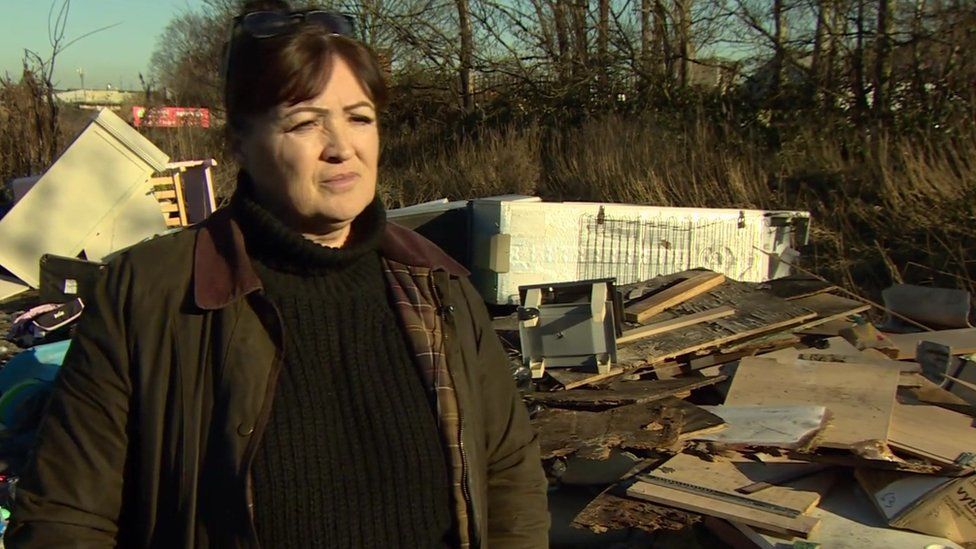Kym Grant, a woman wearing a dark green jacket, stood by fly-tipped rubbish including fridges and bedframes