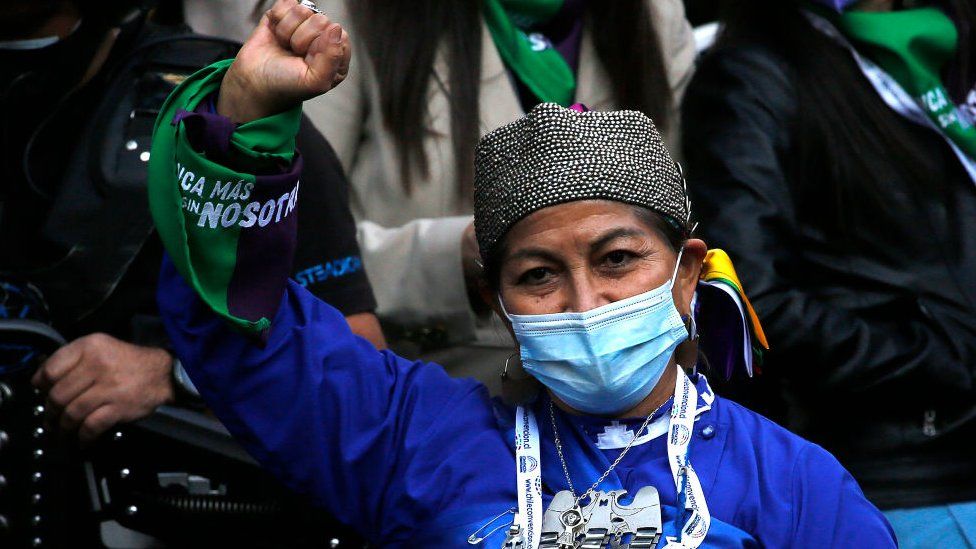 Elisa Loncón after being elected, wearing a face mask due to Covid regulations, dressed with traditional Mapuche clothes and silver jewellery, and holding her right fist in the air as a symbol of pride for her people.