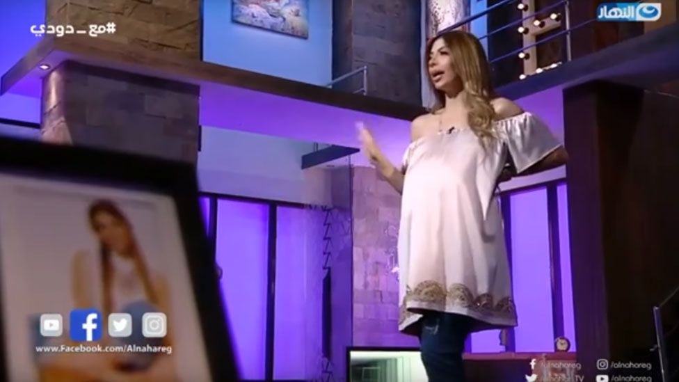 Doaa Salah appears on her show's set dressed to seem pregnant, gesturing towards the camera