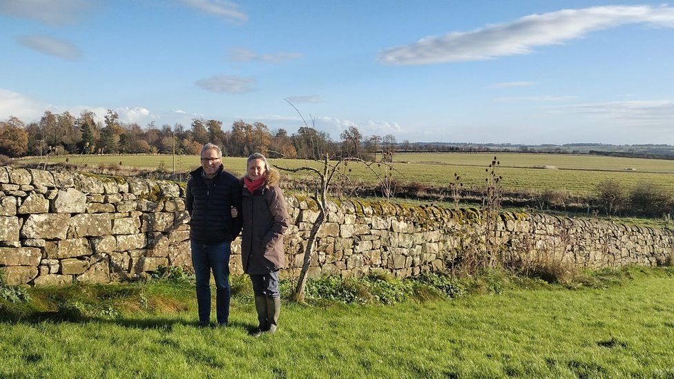Charlotte and Charlie Bennett in front of a stone wall with fields and hills behind them