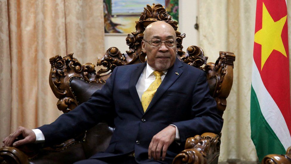 President Desi Bouterse sitting in a wooden chair with a red flag beside him