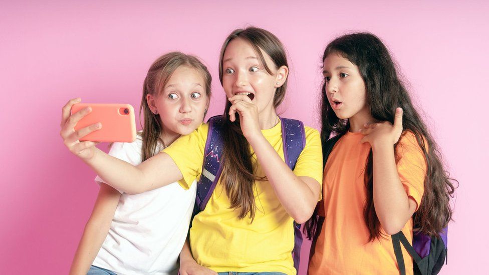 Three teenage girls smiling and looking into a mobile phone on a pink background