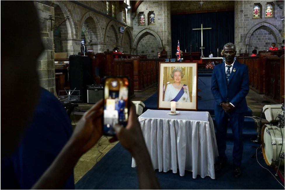 A man posing for a photo in a church standing next to a photo of the Queen. There is a large cross behing him.