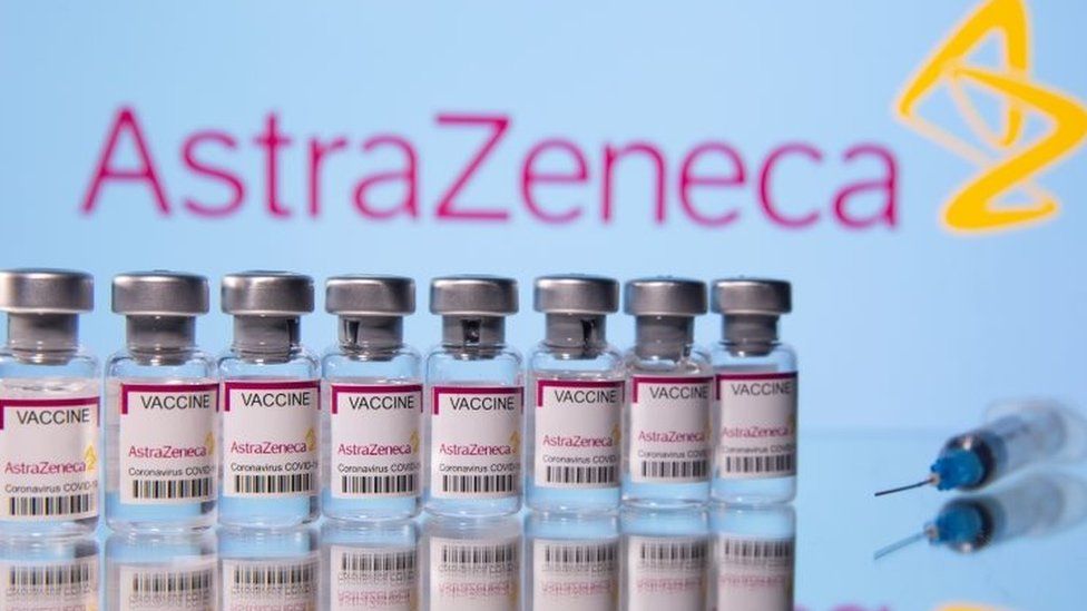 Флаконы с надписью «Astra Zeneca COVID-19 Coronavirus Vaccine» и шприц видны перед отображаемым логотипом AstraZeneca. Файл фото