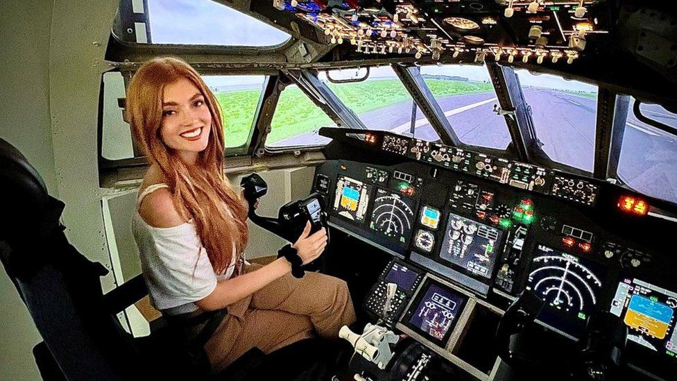 Jessica is sat in the cockpit of a plane holding the controls. She has long ginger hair and is wearing a white top and brown trousers