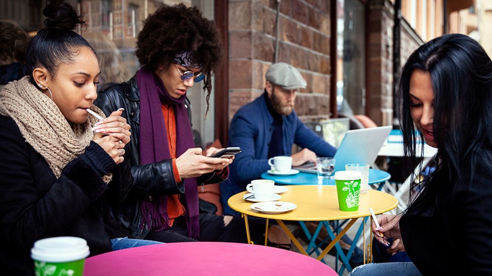 Stock photo shows a group of young people smoking and using mobile and laptop outside a cafe 