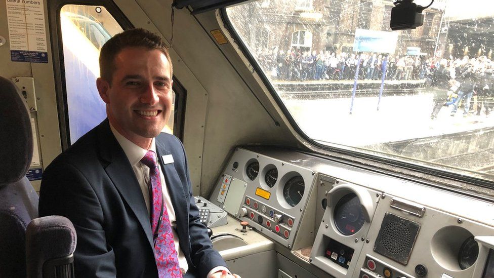 Man with short dark hair and wearing a suit sitting in the driver's seat of a train