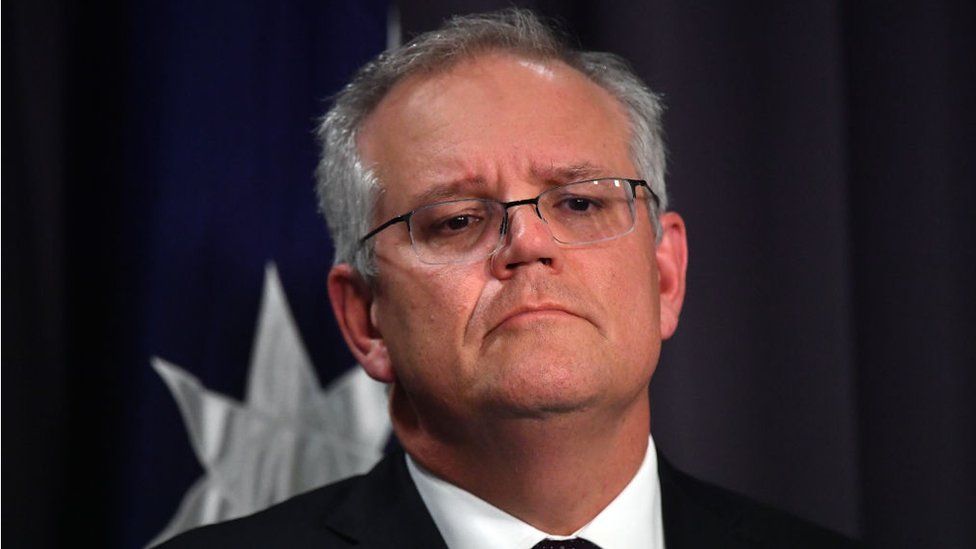 Scott Morrison looks at the camera, with and Australian flag partially visible in the background