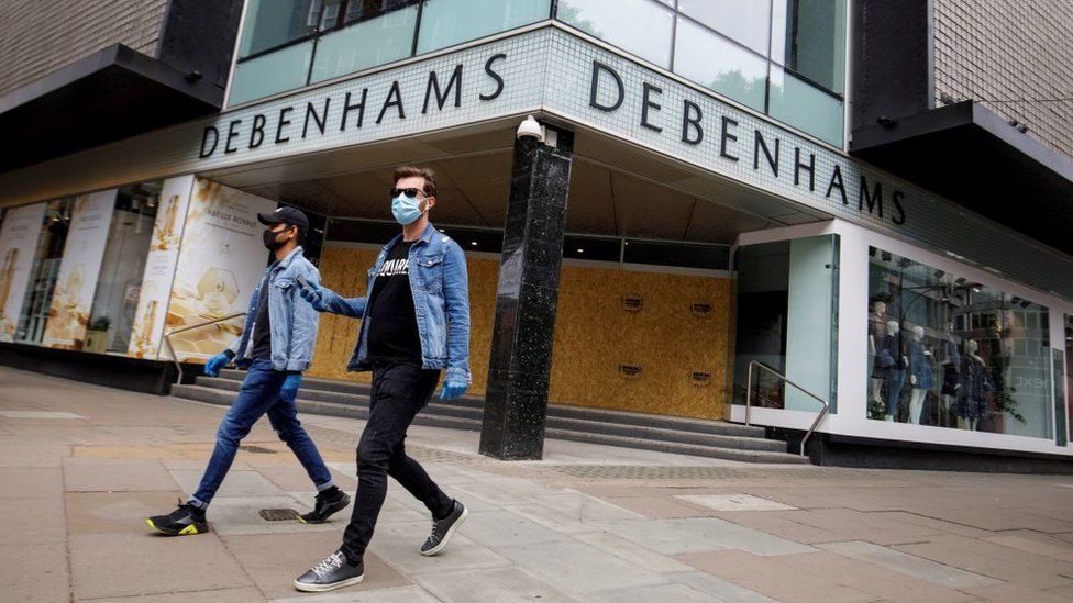 Two people wearing masks walk past a boarded-up Debenhams store in Oxford Street, London