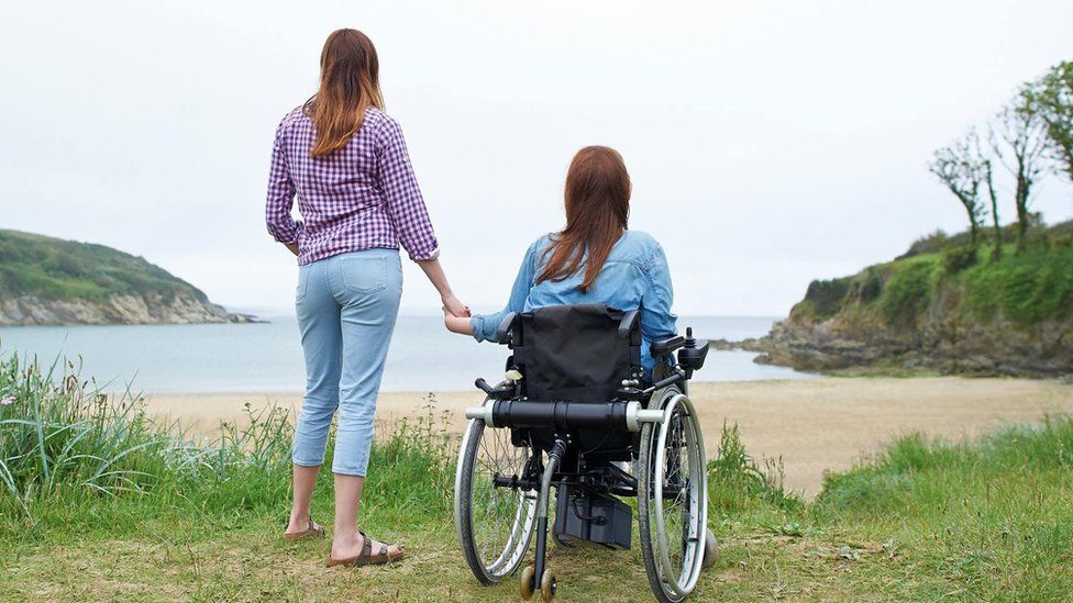 A woman in a wheelchair holding hands with a woman by the coast